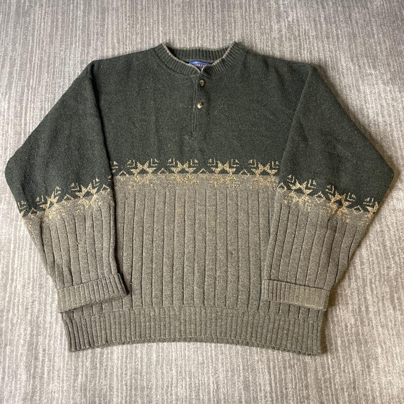 K&ouml;nnte beinhalten: Ein gr&uuml;ner und grauer Pullover mit Rippstrickmuster. Der Pullover hat einen Rundhalsausschnitt mit drei Kn&ouml;pfen und ein dekoratives Muster in einer helleren Farbe &uuml;ber Brust und &Auml;rmeln.