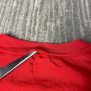 May include: Close-up of a red t-shirt collar with a small tear. Silver scissors are positioned near the tear. The background is a grey, textured surface.