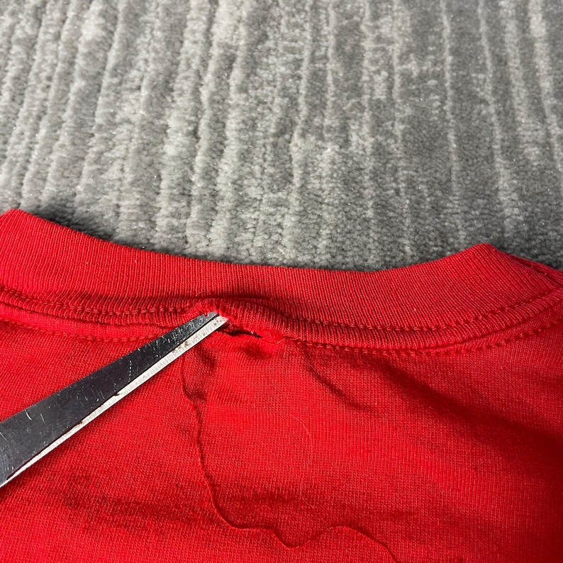 May include: Close-up of a red t-shirt collar with a small tear. Silver scissors are positioned near the tear. The background is a grey, textured surface.