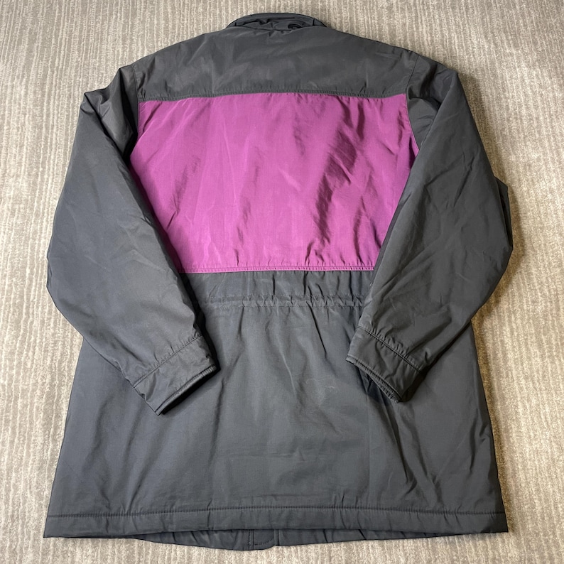K&ouml;nnte beinhalten: Dunkelgraue Jacke mit einem leuchtend lila Einsatz auf dem oberen R&uuml;cken. Die Jacke hat einen Kragen, lange &Auml;rmel und einen Kordelzug in der Taille. Die R&uuml;ckansicht zeigt das Design und die Konstruktion der Jacke.