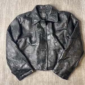 May include: A black leather jacket featuring a patchwork design of stitched leather panels. The jacket has a classic collar, long sleeves, and a front button closure with three black buttons. The interior label reads Genuine Leather.
