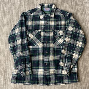 May include: A long-sleeve button-up shirt featuring a plaid pattern in dark green, navy blue, and cream. The shirt includes a pointed collar, two chest pockets with buttoned flaps, and buttoned cuffs. A Pendleton brand label is visible at the neck.