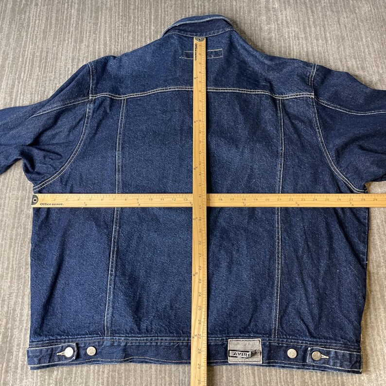 Pode incluir: Uma jaqueta jeans azul escuro com gola, mangas compridas e bainha abotoada. Duas r&eacute;guas de madeira est&atilde;o colocadas sobre as costas da jaqueta, mostrando um comprimento vertical de 61 cm e uma largura horizontal de 61 cm. Uma etiqueta cinza diz AVIREX.