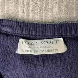Puede incluir: Un su&eacute;ter de punto azul marino oscuro con cuello acanalado. Una etiqueta de tela rectangular blanca est&aacute; cosida en el interior, mostrando el texto verde: LYLE & SCOTT, MADE IN SCOTLAND, TOUCH OF CASHMERE. La textura del tejido es visible.