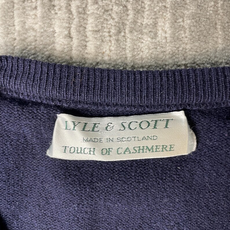 Puede incluir: Un su&eacute;ter de punto azul marino oscuro con cuello acanalado. Una etiqueta de tela rectangular blanca est&aacute; cosida en el interior, mostrando el texto verde: LYLE & SCOTT, MADE IN SCOTLAND, TOUCH OF CASHMERE. La textura del tejido es visible.
