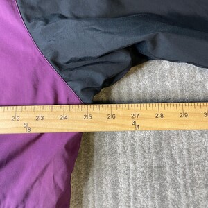 K&ouml;nnte beinhalten: Nahaufnahme einer schwarzen und lila Jacke. Die Jacke hat eine lila Bahn auf der linken Seite und eine schwarze Bahn auf der rechten Seite. Ein Holzlineal liegt &uuml;ber der Jacke und misst ca. 13 cm.