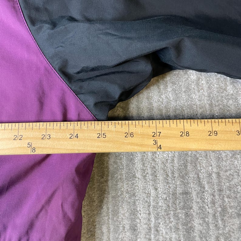 K&ouml;nnte beinhalten: Nahaufnahme einer schwarzen und lila Jacke. Die Jacke hat eine lila Bahn auf der linken Seite und eine schwarze Bahn auf der rechten Seite. Ein Holzlineal liegt &uuml;ber der Jacke und misst ca. 13 cm.