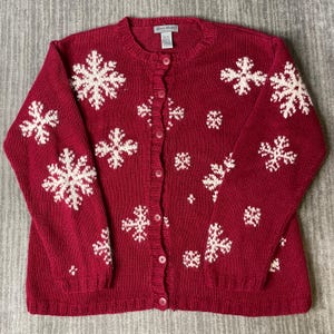 May include: A deep red long-sleeve knit cardigan sweater featuring a repeating white snowflake pattern across the front and sleeves. The garment has a round neckline, a button-down front closure with matching red buttons, and ribbed cuffs.