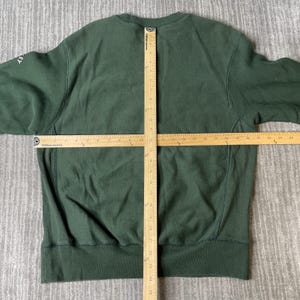 May include: A dark green sweatshirt with a crew neck and long sleeves. The sweatshirt is laid flat on a grey surface, with a ruler placed across the front. The word "key" is visible on the left sleeve.