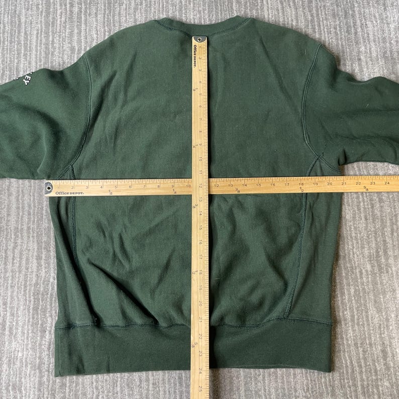 May include: A dark green sweatshirt with a crew neck and long sleeves. The sweatshirt is laid flat on a grey surface, with a ruler placed across the front. The word "key" is visible on the left sleeve.
