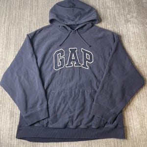 May include: A navy blue long-sleeve hooded sweatshirt featuring the word GAP in large, dark blue embroidered letters with a white outline across the chest. The garment includes a front kangaroo pocket, a drawstring hood, and ribbed cuffs.