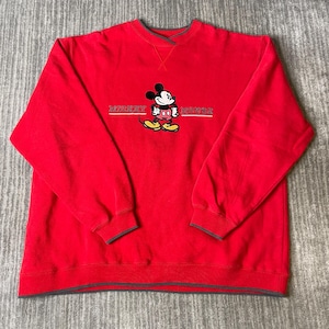Puede incluir: Una sudadera roja de manga larga con cuello redondo y ribete gris oscuro. El centro del pecho presenta un gr&aacute;fico bordado de Mickey Mouse con el texto "MICKEY MOUSE" en letras blancas a ambos lados del personaje.