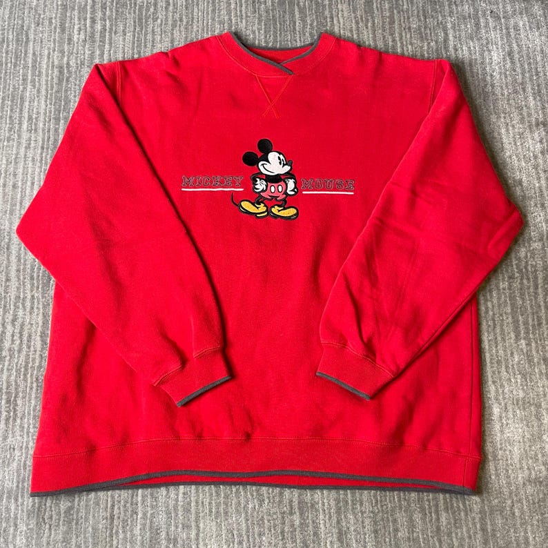 Puede incluir: Una sudadera roja de manga larga con cuello redondo y ribete gris oscuro. El centro del pecho presenta un gr&aacute;fico bordado de Mickey Mouse con el texto "MICKEY MOUSE" en letras blancas a ambos lados del personaje.
