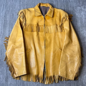 May include: A mustard yellow leather jacket featuring a full-length front zipper, a standard collar, and long sleeves. The garment is decorated with extensive fringe detailing across the chest, shoulders, sleeves, and along the bottom hemline.