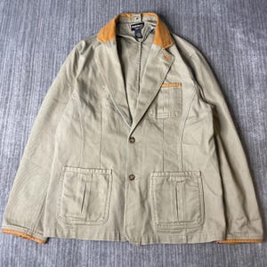 Peut inclure: Un blazer beige avec un col et des poignets de couleur jaune moutarde contrastante. La veste présente deux boutons sur le devant, une poche poitrine et deux grandes poches plaquées. Une étiquette Guide Series est visible au niveau du col.
