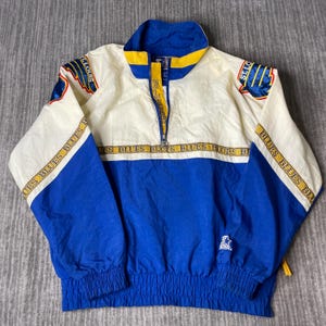 May include: A vintage St. Louis Blues Starter jacket in blue, white, and yellow. The jacket features a half-zip closure, elastic cuffs, and a drawstring hem. The team logo is on the sleeves. The text "BLUES" is printed across the chest.