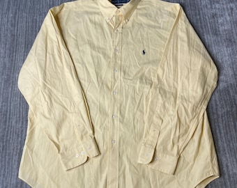 Pode incluir: Uma camisa de manga comprida abotoada em amarelo claro. A camisa apresenta uma gola abotoada, uma carcela frontal com botões e um pequeno logótipo bordado em azul escuro de uma pessoa num cavalo na zona esquerda do peito.