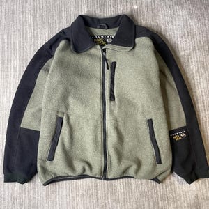 May include: A sage green and black fleece jacket featuring a full-length front zipper, a high collar, and black panels on the sleeves and shoulders. The jacket includes two zippered hand pockets, one zippered chest pocket, and Mountain Hardwear branding.