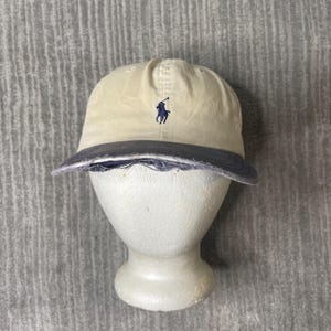 May include: A cream-colored baseball cap featuring a dark blue embroidered polo player logo on the front panel. The cap has a dark blue brim with a distressed, frayed edge along the front, showing a worn texture and visible stitching details.