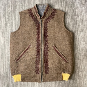 May include: A brown sleeveless vest featuring a full-length front zipper, a high collar, and dark red decorative embroidery along the neckline and zipper. The garment includes two slanted front pockets with matching embroidery and yellow ribbed trim.