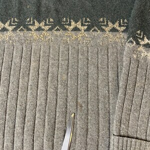 K&ouml;nnte beinhalten: Nahaufnahme eines grau-gr&uuml;nen Pullovers mit gerippter Struktur. Der obere Teil hat ein dekoratives Muster in einer helleren Beigefarbe. Der Pullover hat eine Tasche auf der rechten Seite. Eine Schere befindet sich unten in der Mitte.