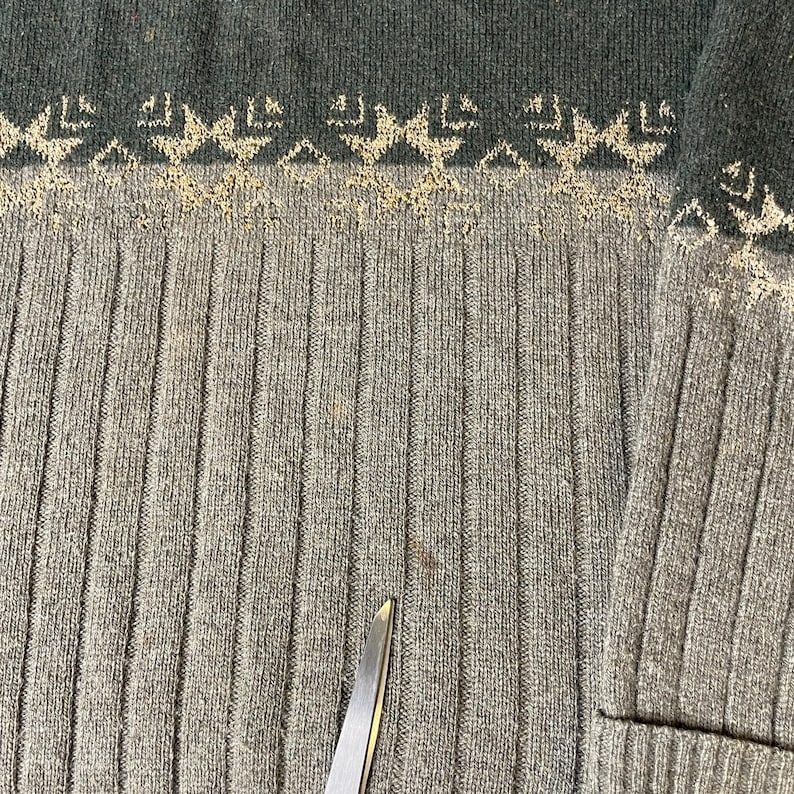 K&ouml;nnte beinhalten: Nahaufnahme eines grau-gr&uuml;nen Pullovers mit gerippter Struktur. Der obere Teil hat ein dekoratives Muster in einer helleren Beigefarbe. Der Pullover hat eine Tasche auf der rechten Seite. Eine Schere befindet sich unten in der Mitte.