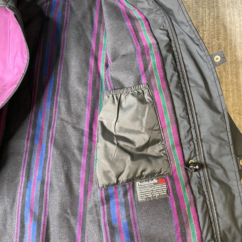 K&ouml;nnte beinhalten: Schwarze Jacke mit lila Futter und gestreiftem Innenfutter in Lila-, Blau- und Gr&uuml;nt&ouml;nen. Die Jacke hat eine schwarze Tasche und ein Etikett mit der Aufschrift "Thinsulate 3M".