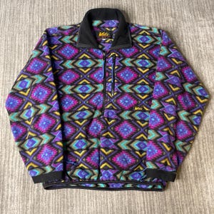 May include: A colorful fleece jacket with a geometric pattern in shades of purple, teal, yellow, and black. The jacket has a black collar, cuffs, and a zipper closure. The REI logo is visible on the collar and chest.