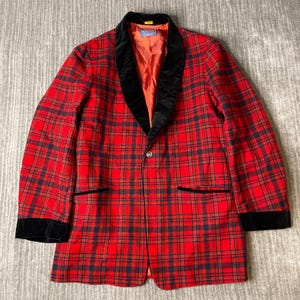 May include: A red and black plaid blazer featuring a black velvet shawl collar, black velvet cuffs, and black velvet trim on the two front pockets. The jacket has a single front button closure and a solid red interior lining with a visible brand label.