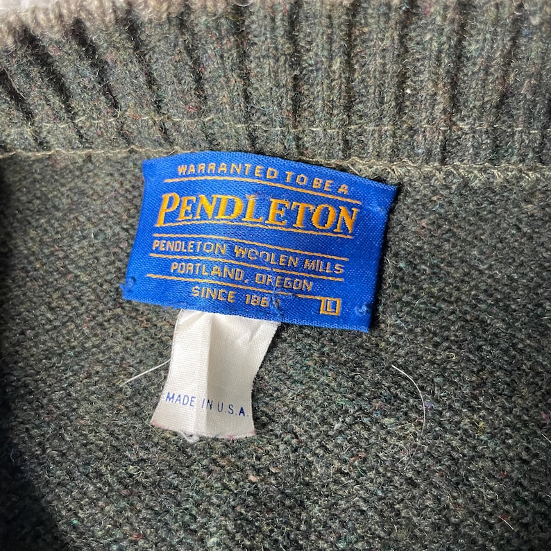 K&ouml;nnte beinhalten: Nahaufnahme eines gr&uuml;nen Wollpullovers mit einem blauen Etikett mit der Aufschrift "PENDLETON" und "MADE IN U.S.A.". Das Etikett besagt auch "PENDLETON WOOLEN MILLS PORTLAND, OREGON SINCE 1863".