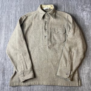May include: A long-sleeve pullover shirt in a light tan, textured fabric. It features a pointed collar, a partial button-down placket with four buttons, and a single chest pocket with a buttoned flap. The cuffs also include button closures.