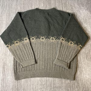 K&ouml;nnte beinhalten: Ein zweifarbiges Sweatshirt in Gr&uuml;n- und Graut&ouml;nen. Der obere Teil ist ein dunkleres Gr&uuml;n, w&auml;hrend der untere Teil ein helleres Grau ist. Ein dekoratives Muster in Beige ziert die &Auml;rmel und die Brust. Das Sweatshirt hat eine gerippte Textur.