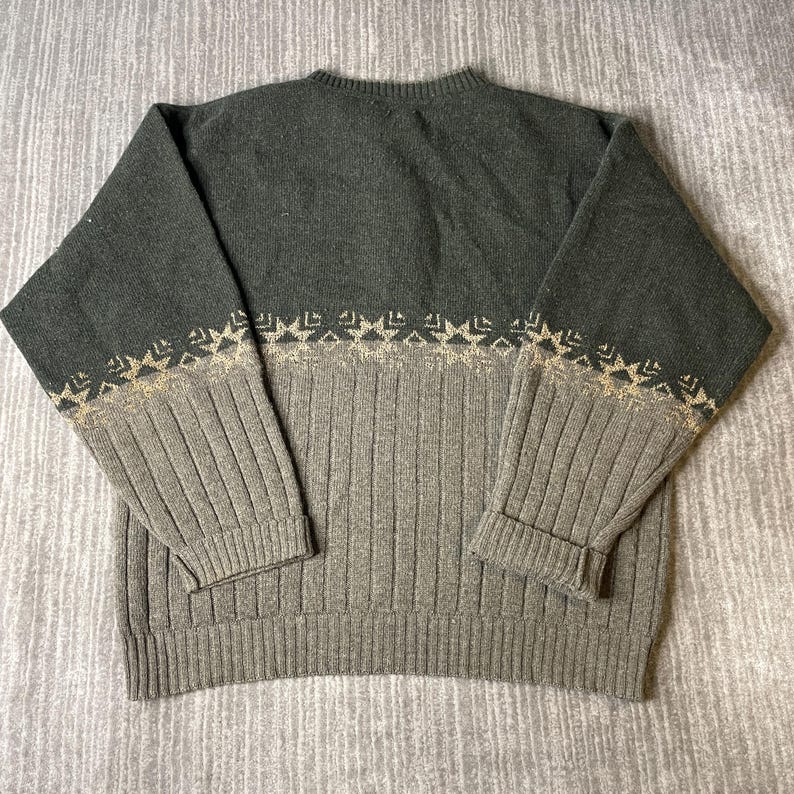 K&ouml;nnte beinhalten: Ein zweifarbiges Sweatshirt in Gr&uuml;n- und Graut&ouml;nen. Der obere Teil ist ein dunkleres Gr&uuml;n, w&auml;hrend der untere Teil ein helleres Grau ist. Ein dekoratives Muster in Beige ziert die &Auml;rmel und die Brust. Das Sweatshirt hat eine gerippte Textur.