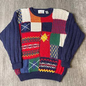 May include: A long-sleeve crewneck sweater featuring a multi-colored patchwork design. The garment displays various knit patterns including cable, ribbed, and textured squares in shades of navy blue, red, cream, green, and yellow. The Arlanda brand label is visible at the inner neckline.