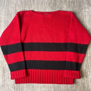 May include: A long-sleeved knit sweater featuring a bold horizontal striped pattern in vibrant red and deep black. The garment has a classic crew neckline, ribbed cuffs at the wrists, and a wide ribbed hemline, constructed from a thick yarn material.