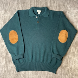 May include: A dark teal long-sleeve knit sweater featuring a polo-style collar with two buttons at the neckline. The garment includes tan-colored oval-shaped elbow patches on each sleeve, a ribbed hem, and ribbed cuffs. A small white label is at the neck.