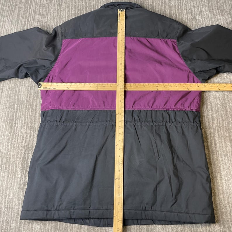 K&ouml;nnte beinhalten: Eine schwarze und lila Jacke mit Kordelzug in der Taille. Die Jacke hat einen lila Einsatz auf Brust und R&uuml;cken, mit schwarzen Eins&auml;tzen an den &Auml;rmeln und am unteren K&ouml;rper. Die Jacke liegt flach auf einer neutralen Oberfl&auml;che.