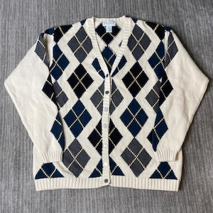 May include: Cream-colored argyle cardigan sweater with a V-neck and button closure. The sweater features a pattern of navy blue, black, gray, and tan diamond shapes. The brand name "IZOD" is visible on the label.