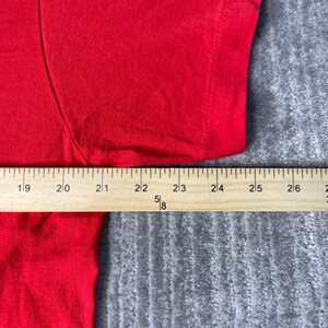 May include: A close-up of a red t-shirt with a wooden ruler placed across the fabric. The ruler measures from 48 to 66 cm. The shirt is on a grey surface.