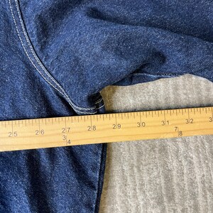 Pode incluir: Uma pe&ccedil;a de vestu&aacute;rio em ganga azul escura com costuras brancas vis&iacute;veis ao longo das jun&ccedil;&otilde;es. Uma r&eacute;gua de madeira est&aacute; colocada sobre o tecido, mostrando uma medida de 63,5 cm a mais de 81 cm, indicando a largura do artigo.