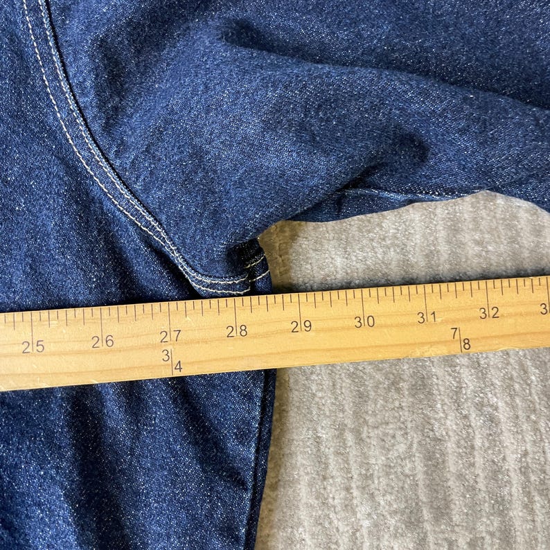Pode incluir: Uma pe&ccedil;a de vestu&aacute;rio em ganga azul escura com costuras brancas vis&iacute;veis ao longo das jun&ccedil;&otilde;es. Uma r&eacute;gua de madeira est&aacute; colocada sobre o tecido, mostrando uma medida de 63,5 cm a mais de 81 cm, indicando a largura do artigo.
