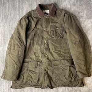May include: An olive green field jacket featuring a brown corduroy collar, a button-front closure, and long sleeves. The garment includes one chest pocket with a buttoned flap and two large lower patch pockets. A label is visible at the neckline.