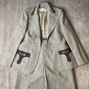 May include: A grey wool-blend blazer and skirt set featuring dark brown suede-like pocket flaps with silver-tone metal horsebit hardware. The blazer has a notched collar, two front buttons, and a label reading Lilli Ann San Francisco.