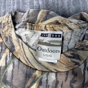 以下が含まれることがあります： 「JERZEES Outdoors L/G/G」と書かれたタグが付いた迷彩柄のTシャツ。シャツは茶色、タン、緑色のブレンドで、クルーネックです。タグには風景グラフィックが描かれています。