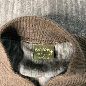 May include: Close-up of a camouflage pattern shirt collar with a brown ribbed knit neckline. A green rectangular tag reads "SPARTAN REALTREE MADE IN USA". The shirt's fabric features a mix of green, brown, and tan colors.