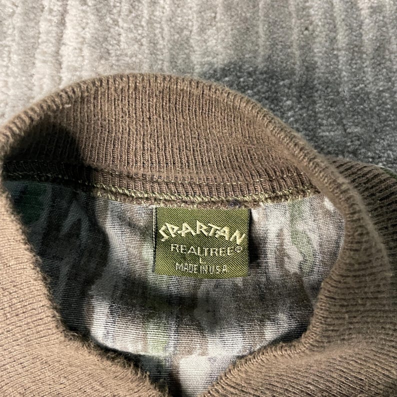 May include: Close-up of a camouflage pattern shirt collar with a brown ribbed knit neckline. A green rectangular tag reads "SPARTAN REALTREE MADE IN USA". The shirt's fabric features a mix of green, brown, and tan colors.
