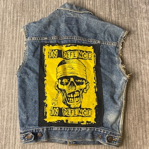 May include: A blue denim vest with frayed armholes featuring a large rectangular patch on the back. The patch has a yellow background with black text reading IN DEFENCE at the top and bottom, and a graphic of a skull wearing a bandage.