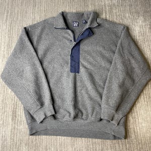 May include: A gray fleece pullover with a navy blue accent panel and collar. The pullover has a zipper closure and the Gap logo on the collar. The cuffs and hem are ribbed.