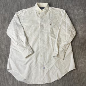 May include: A long-sleeved, button-down white shirt with a button-down collar. The shirt features a small embroidered logo on the chest and buttoned cuffs. The shirt is made of a lightweight fabric.