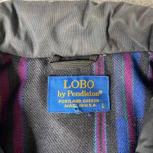 K&ouml;nnte beinhalten: Nahaufnahme eines Jackenkragens mit einem blauen Etikett mit der Aufschrift "LOBO by Pendleton" und "PORTLAND, OREGON MADE IN U.S.A.". Das Innenfutter hat ein farbenfrohes Streifenmuster. Die Jacke ist schwarz.